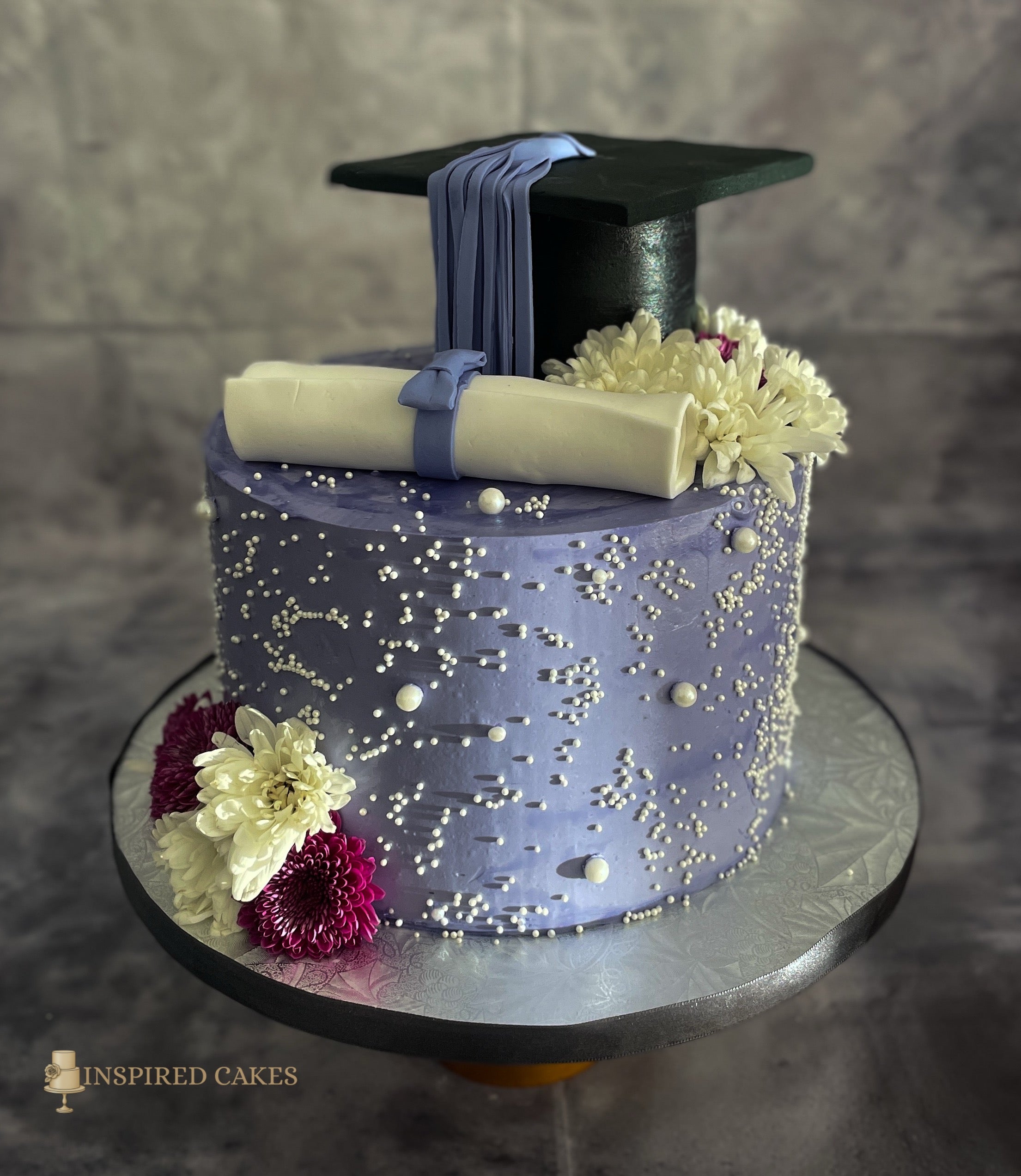 Graduation-themed cake with a cap, diploma, and flowers on a gray background.